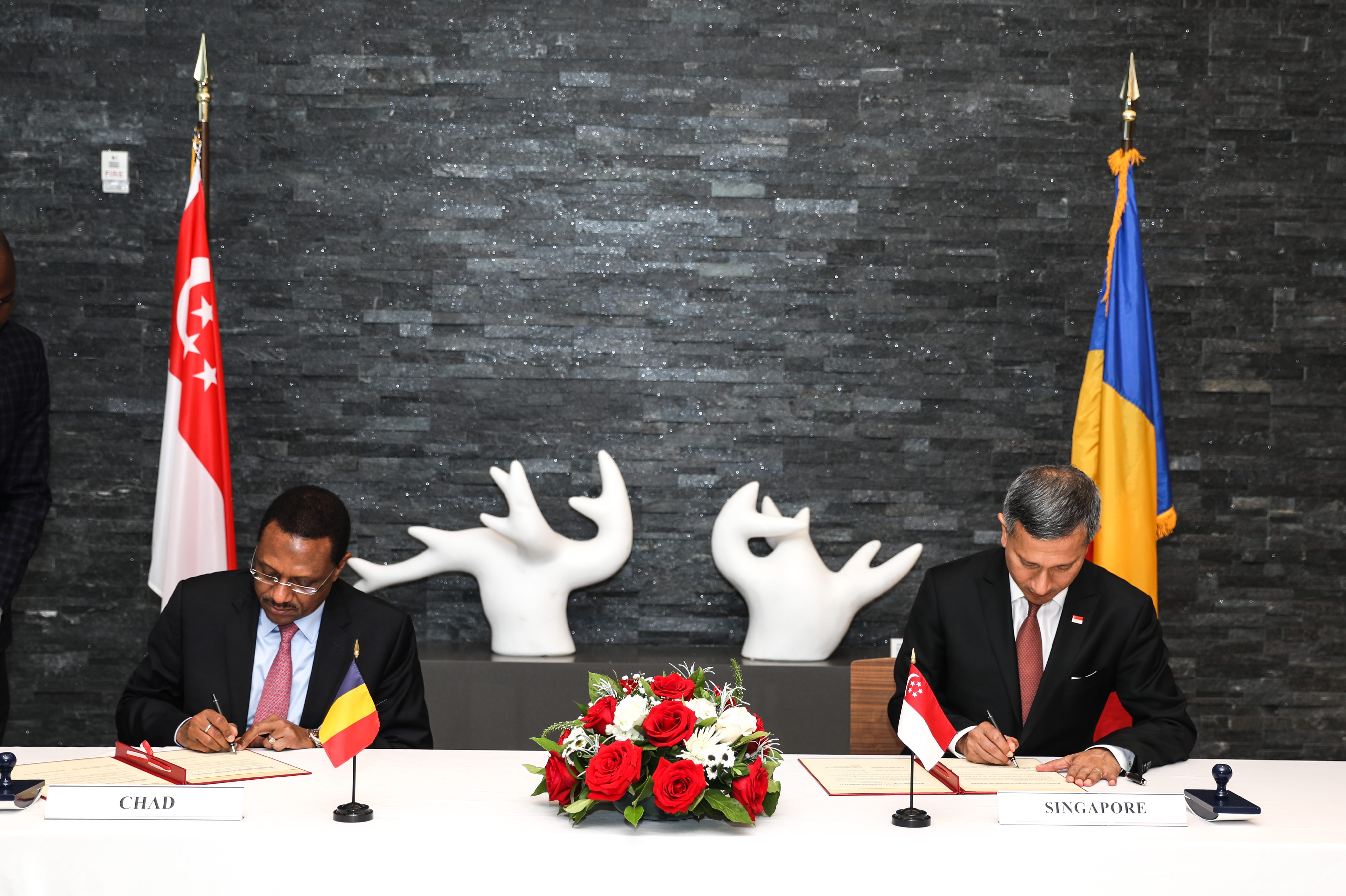 Two men signing agreements for Chad and Singapore, with flags, rose centerpiece, and modern art backdrop.